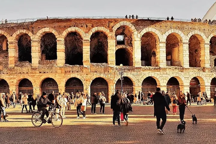 The pink stone arches of the Arena di Verona on Piazza Bra in warm evening light, with people and a cyclist passing
